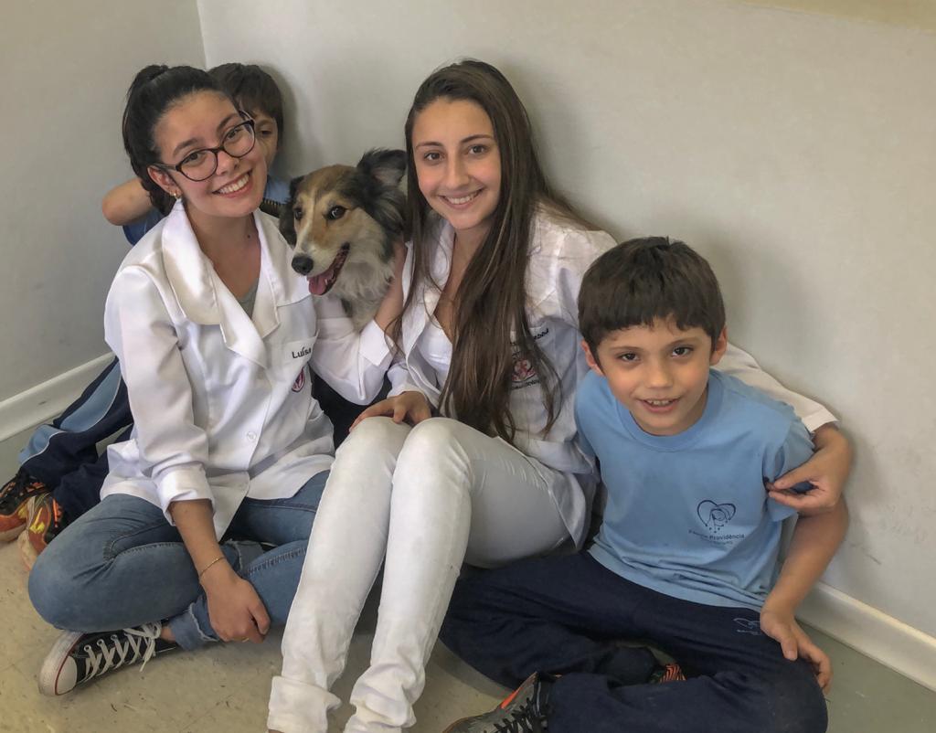 Duas estudantes de jaleco branco agachadas com uma criança e um cachorro