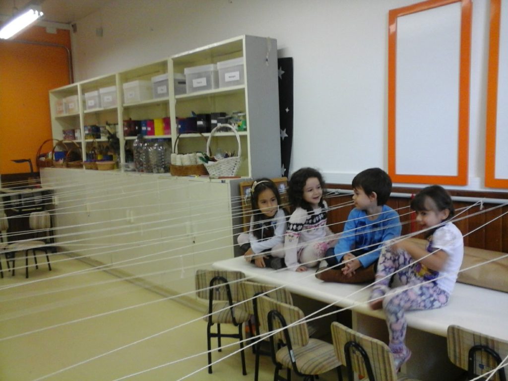 Na foto há quatro crianças sentadas em uma mesa, brincando com fios brancos. Os fios estão amarrados horizontalmente para as crianças