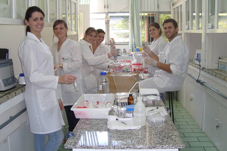 Seis pessoas vestindo jaleco branco atrás de uma mesa com equipamentos de química