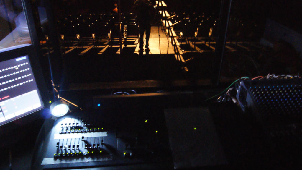 Mesa de operação de som em primeiro plano e salão do teatro ao fundo com diversas cadeiras e luz difusa