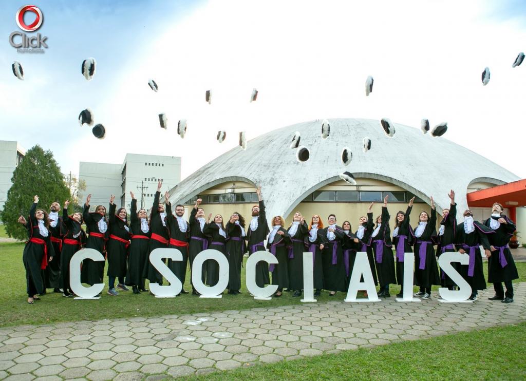 Alunos com becas atras de um letreiro representando as ciencias sociais, jogam os capelos para cima, comemorando a formatura