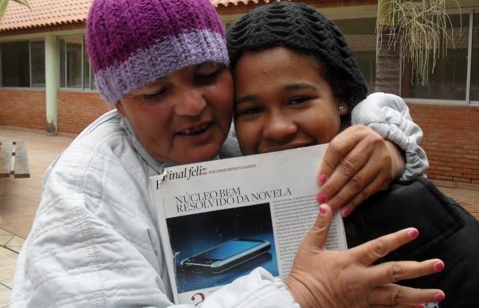 Uma senhora segurando uma revista, abraça uma aluna durante uma visita ao Lar das vovozinhas