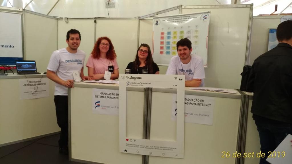 quatro alunos brancos, sendo dois homens e duas mulheres, atrás de um balcão branco sorrindo para foto durante o Descubra 2019