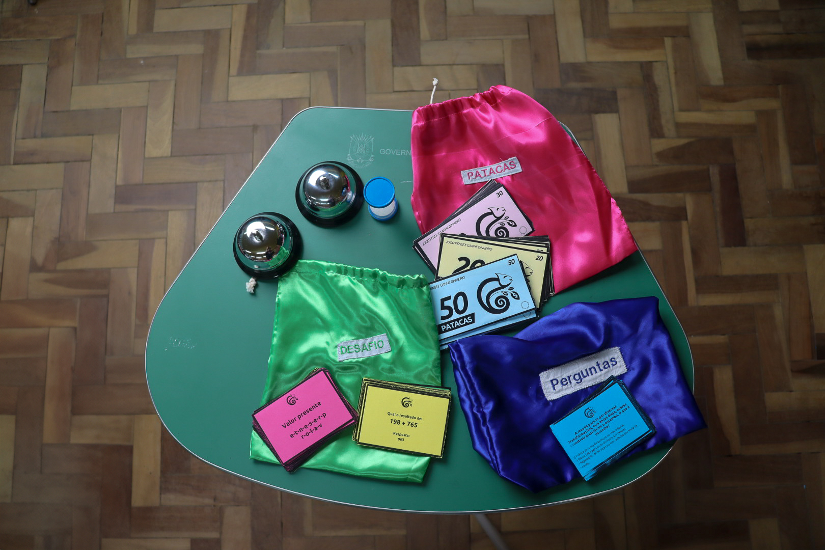 Jogo Pense e Ganhe Dinheiro sobre mesa escolar. Sobre a mesa, duas sinetas em tom prata e uma ampulheta de plástico. Também tem cédulas de dinheiro pataca, do jogo, sobre sacos de tecido coloridos em tons verde, rosa e azul.