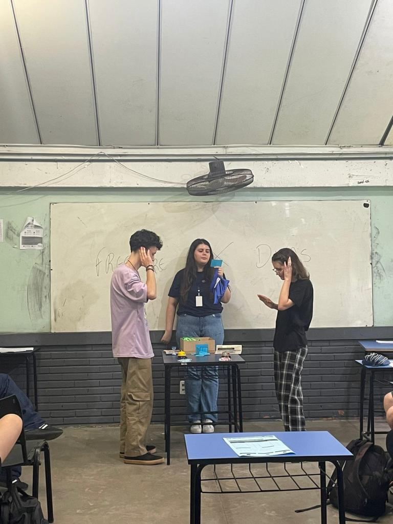 Fotografia de três estudantes em sala de aula. Há uma mesa entre dois deles, que se olham e estão com uma das mãos atrás da orelha e a outra apontada sobre a mesa. Atrás da classe escolar, uma mulher em pé lê uma carta. Sobre a mesa, há uma sineta, ampulheta e elementos em papeis coloridos.