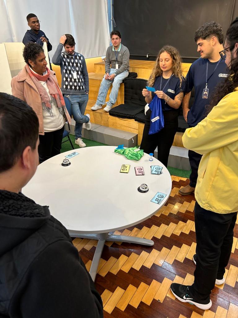 Cerca de oito pessoas estão em pé, espalhadas em um ambiente de sala de aula. No meio da foto, há uma mesa redonda, em que estão elementos de um jogo: cartas coloridas, sineta e ampulheta. Uma mulher segura um saco de tecido e lê uma das cartinhas.