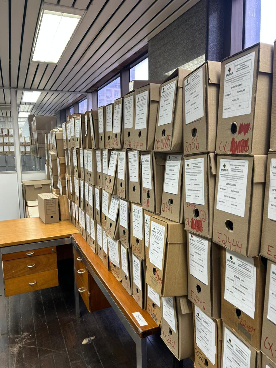 Sala de arquivo com diversas caixas de documentos de papel empilhadas sobre mesas e estantes. As caixas possuem etiquetas de identificação e anotações manuscritas. Ao fundo, há armários, janelas e iluminação de teto.