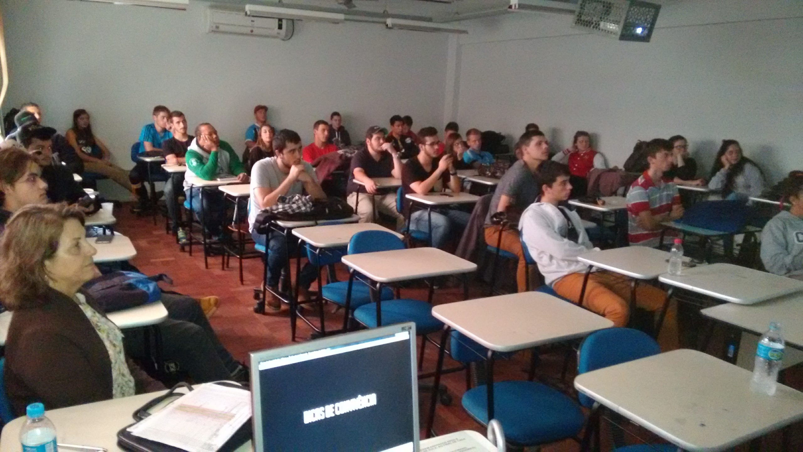 Descri&ccedil;&atilde;o da fotografia: Alunos sentados nas cadeiras em fileiras, alguns debru&ccedil;ados nas classes. Todos olham na mesma dire&ccedil;&atilde;o.
