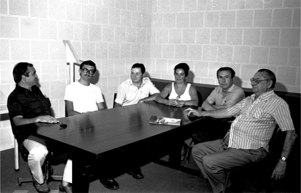  Fotografia em preto e branco, em plano geral, de cinco homens e uma mulher sentados ao redor de uma mesa retangular escura. Sobre a mesa, um óculos de sol e um folheto. Ao fundo, parede de revestimento acústico branco e um púlpito.