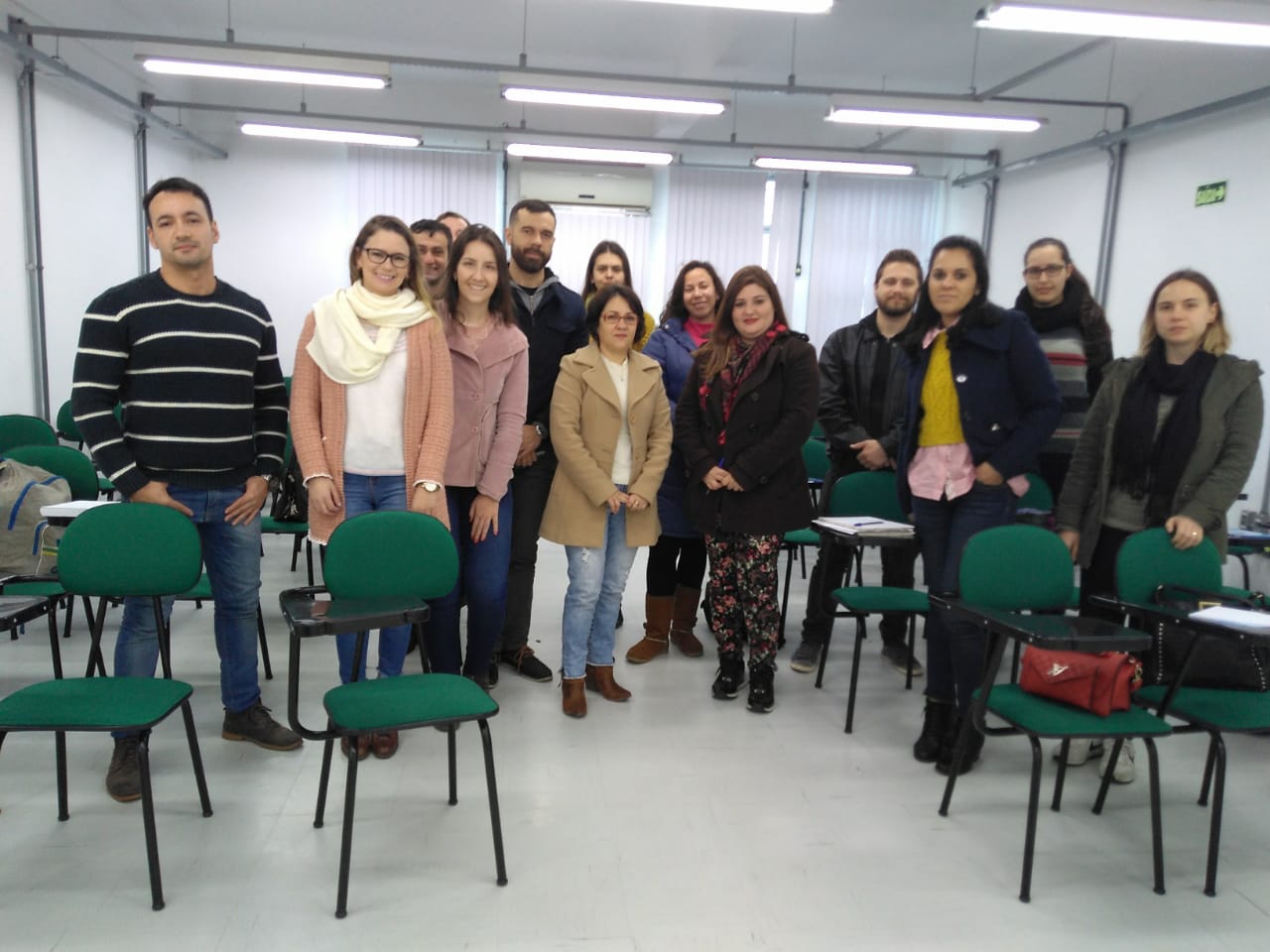 Foto mostra 14 servidores em pé, lado a lado, em uma sala de aula com cadeiras verdes