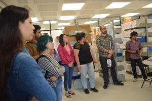 Imagem colorida, na horizontal. sete pessoas em uma sala. Ao fundo, três prateleiras com arquivos.