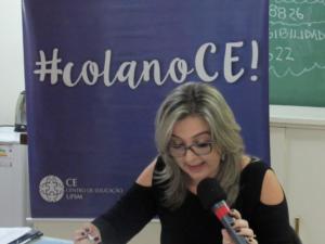 Foto horizontal. Uma professora com um microfone na mão e braços apoiados em uma mesa branca. Atrás dela há um banner escrito "#colanoCE"