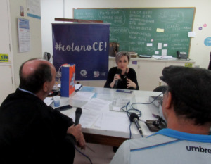 Foto horizontal. Uma professora sentado com um microfone na mãe. Em sua frente, há dois homens, também segurando microfones. Entre eles há uma mesa branca.