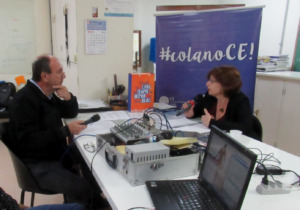 Foto horizontal. Uma professora está sentada em uma cadeira, segurando um microfone. Em sua frente há um jornalista segurando um microfone.  Entre eles há uma mesa branca com equipamentos de gravação e um notebook.
