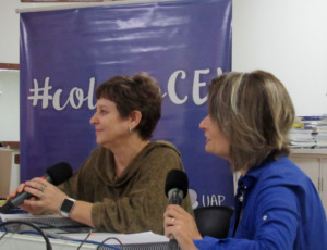 Foto horizontal. Duas professoras estão sentadas lado a lado sendo entrevistadas. As duas seguram um microfone. 