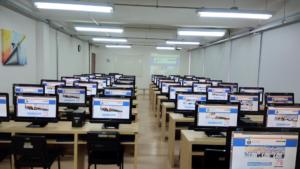Duas fotografias horizontais tiradas da frente para o fundo da sala, de um laboratório de informática. Há três computadores - na cor preta, por mesa - da cor bege, enfileirados do início ao fim da foto. Na parte superior, aparecem lâmpadas acesas na cor branca, também enfileiradas, e na parede do lado esquerdo há um quadro colorido pendurado. 