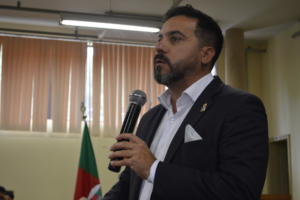 Professor Flavi Ferreira Lisboa Filho, Pró-reitor de Extensão da UFSM, fala ao microfone. Homem branco de cabelos castanhos com barba castanha. Veste terno azul com camisa branca. Ao fundo, janela e bandeira do Rio Grande do Sul.