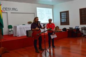 Da esquerda para a direita, homem branco, de cabelos loiros no comprimento até os ombros, veste calça jeans preta e camiseta preta. Ele segura um violão e fala ao microfone que está preso a um pedestal. Ao lado, Prof Sidnei, homem branco de cabelos pretos curtos, veste calça preta e camiseta vermelha. Segura um papel com a mão direita e segura o braço direito, na altura do cotovelo, com a mão esquerda.