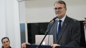 Paulo Afonso Burmann, Reitor da UFSM. Homem branco, com cabelo parcialmente grisalho, vestindo terno cinza, camiseta branca e gravata azul. Ele está falando ao microfone, em frente a um púlpito de cor preta. Em cima do púlpito há um notebook cinza.