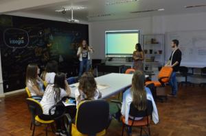 Alunos sentados ao redor de uma mesa assistindo a uma apresentação de três alunos que estão em pé. Os alunos sentados estão de costas para a foto e os alunos em pé estão voltados de frente para a fotografia. O local é a Agência Íntegra do Decom UFSM-FW