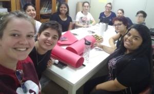 Em uma pequena sala, vê-se ao fundo uma porta fechada, na parede do lado esquerdo um armário com três portas de vidro, através das quais se podem ver muitos livros. No canto esquerdo da sala, uma pequena escrivaninha e sobre ela um computador. No centro, uma grande mesa retangular branca que ocupa quase todo o espaço da sala. Sentadas ao redor da mesa, estão quatro professoras e seis bolsistas que fazem parte da equipe que atua nos berçários da Unidade de Educação Infantil Ipê Amarelo. A maioria das pessoas da equipe, estão sorrindo. No centro da mesa está um prato com biscoitos e um rolo de papel toalha, também estão sobre a mesa uma garrafa térmica de aço inoxidável com tampa preta, algumas folhas de papel ofício, duas folhas de cartolina rosa enroladas e outra dobrada. Ainda sobre a mesa estão duas agendas, três aparelhos de celular, uma garrafa de água e uma squeeze plástica transparente com tampa preta.